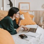woman drinking from a cup on her computer on a bed