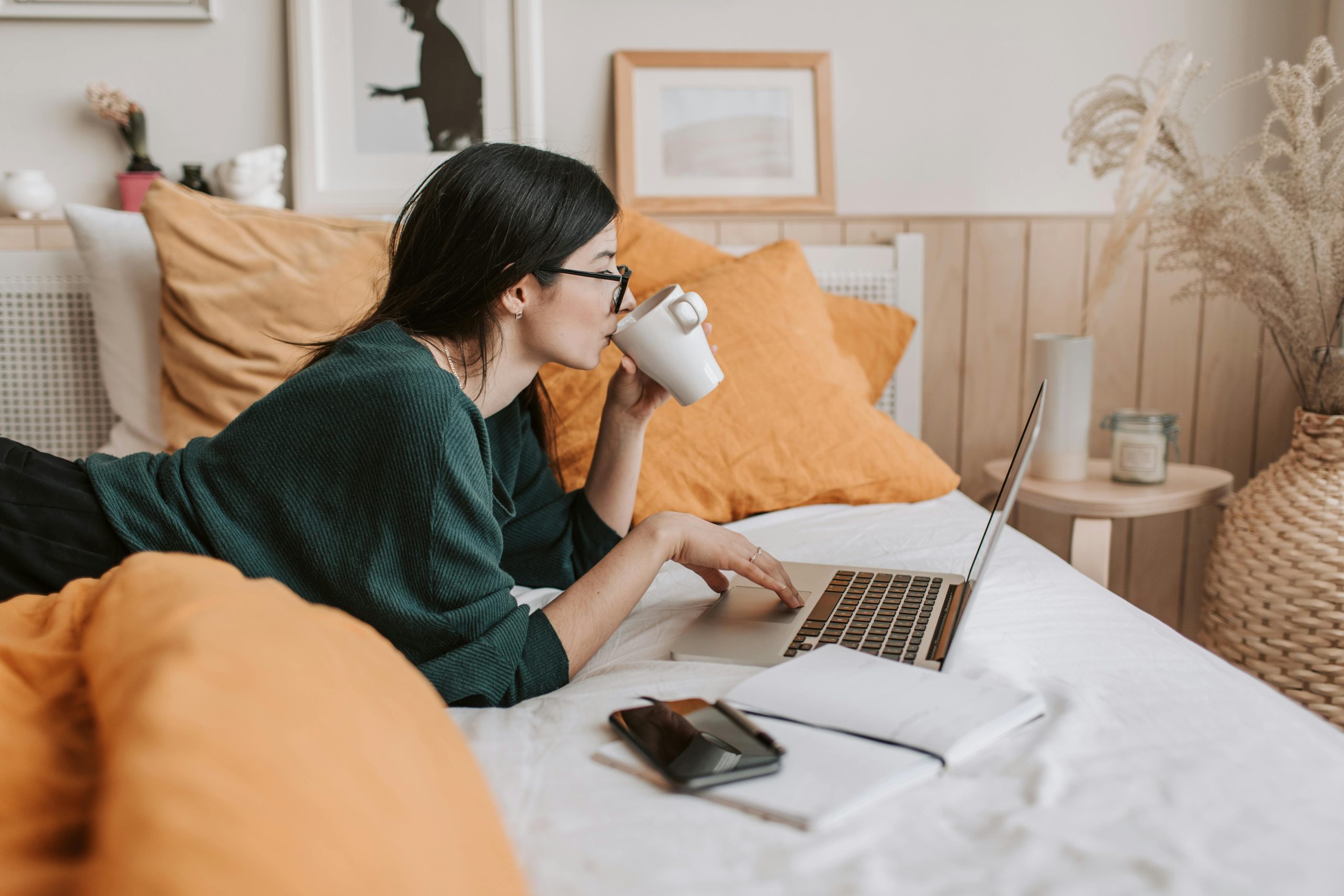 woman drinking from a cup on her computer on a bed